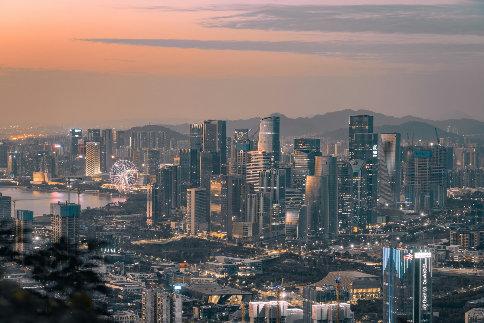 The skyline of Qianhai, Shenzhen, at dusk, with a ferris wheel and Qianhai Bay at left and construction cranes among the towers